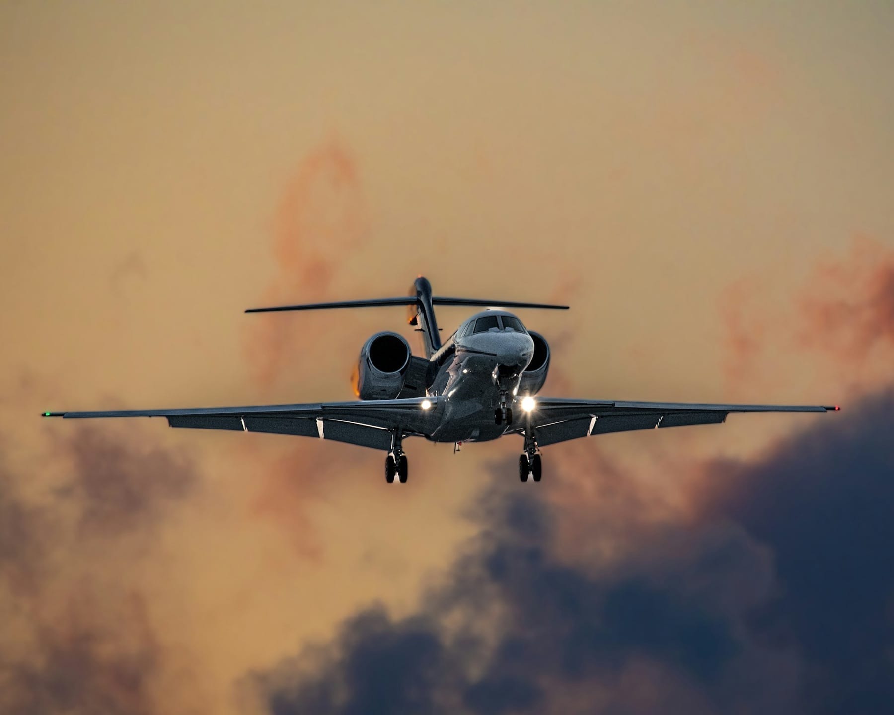 Business jet on final approach with landing gear down and landing lights on, against a sunset sky