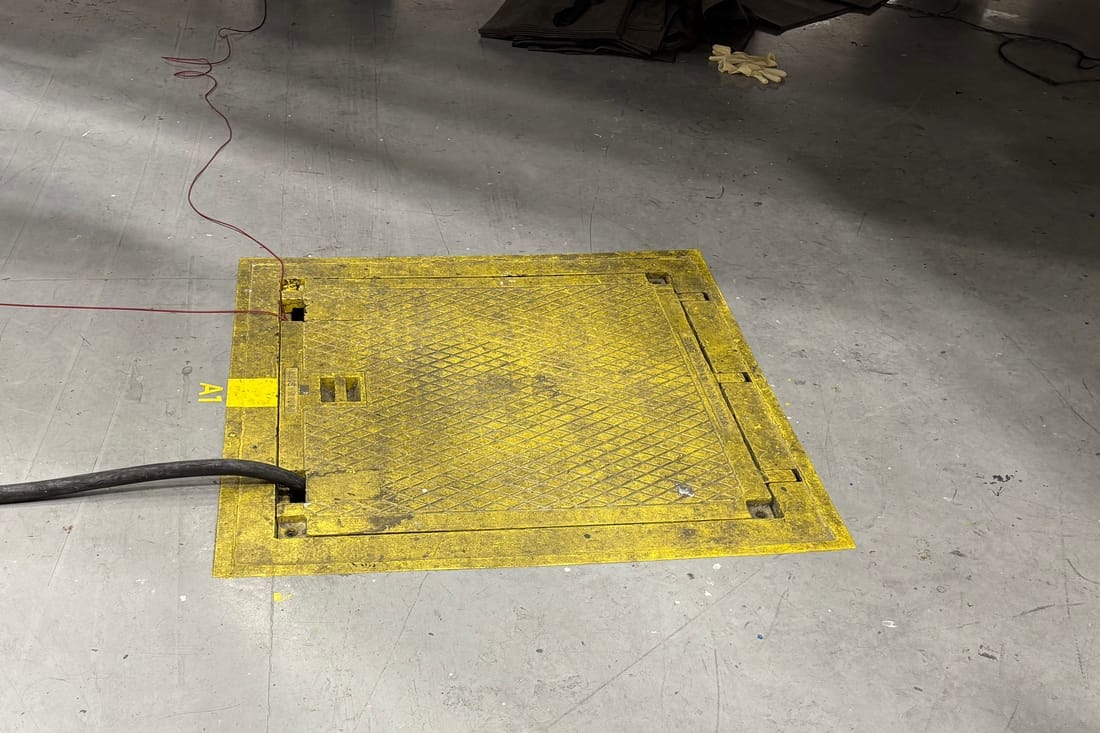 A utility pit set into a hangar floor, its yellow cover flush with the concrete, a power cable running out of one edge and a painted position marking beside it