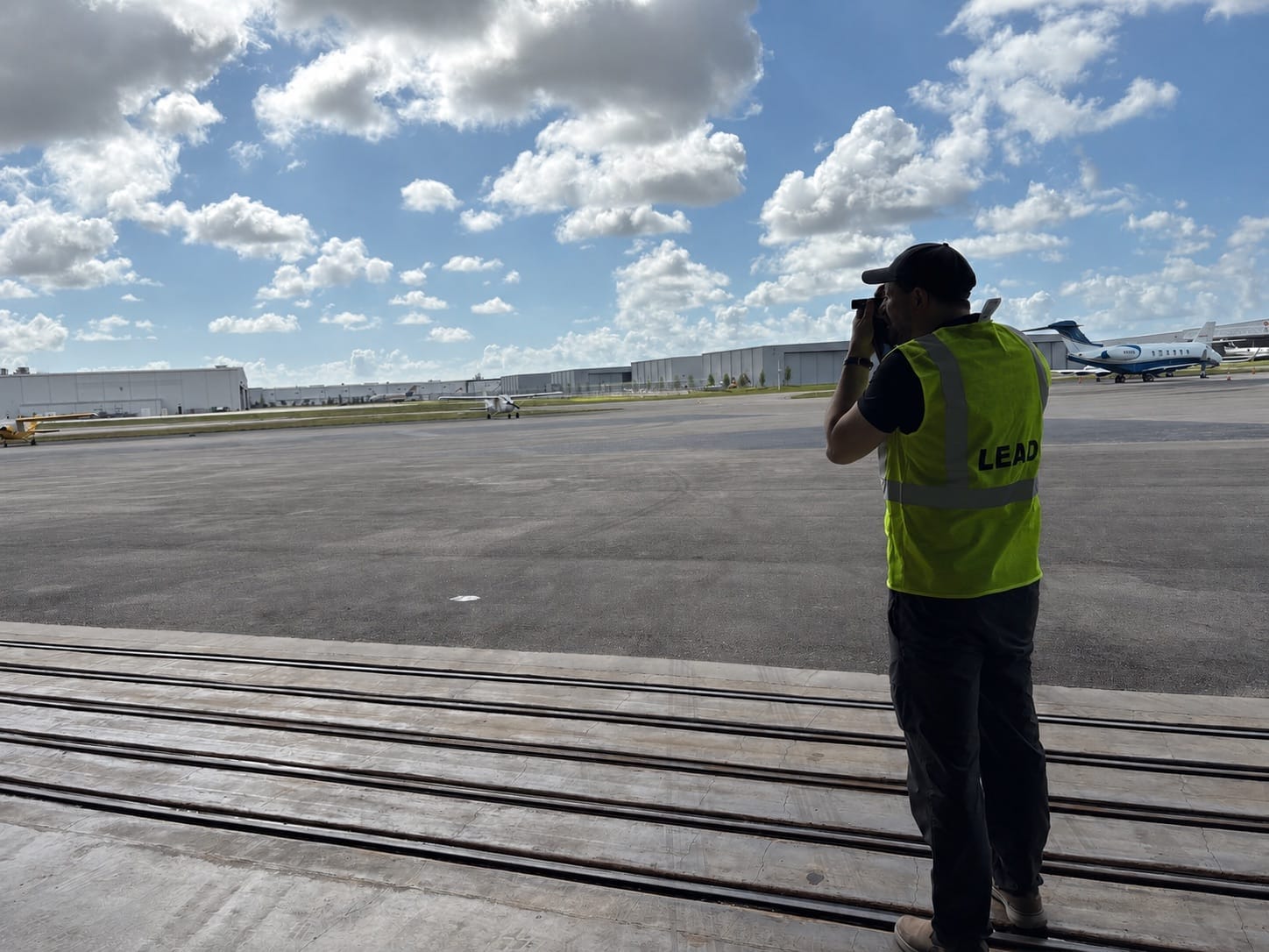 An AirPlx team member in a high-visibility vest standing on the hangar door rail, sighting a laser rangefinder across the ramp, with business jets and hangars beyond